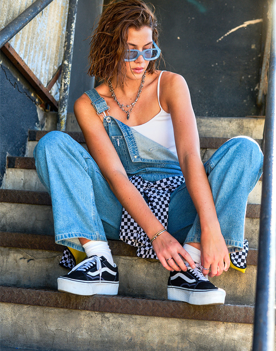 A woman laces up her Platform Old Skool on a concrete staircase wearing a white Drew Rib Tank Top and denim blue Sirelle Overalls.