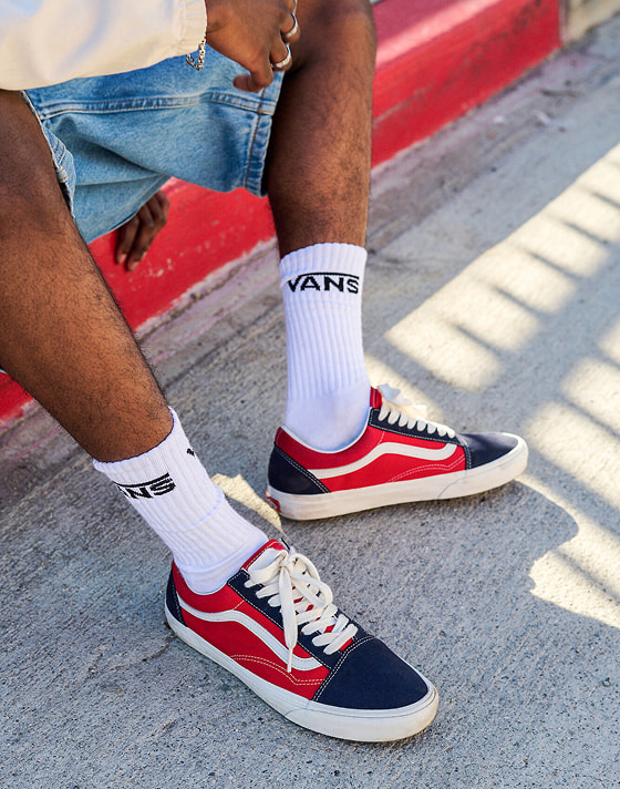 A close-up of the a man wearing red and navy Old Skool paired with blue denim shorts sitting on a curb.