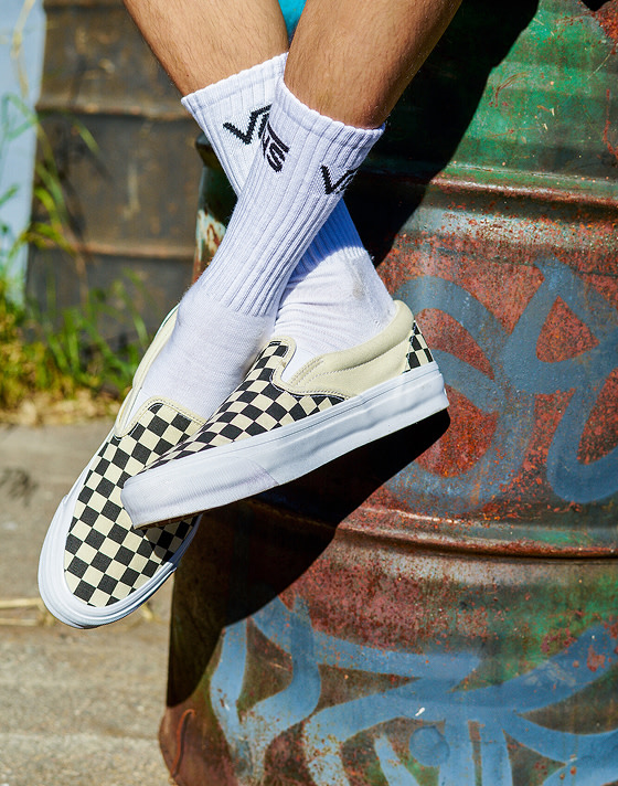 A person crosses their legs while sitting on an oil drum wearing white Vans crew socks and black and white Checkerboard Classic Slip-On.