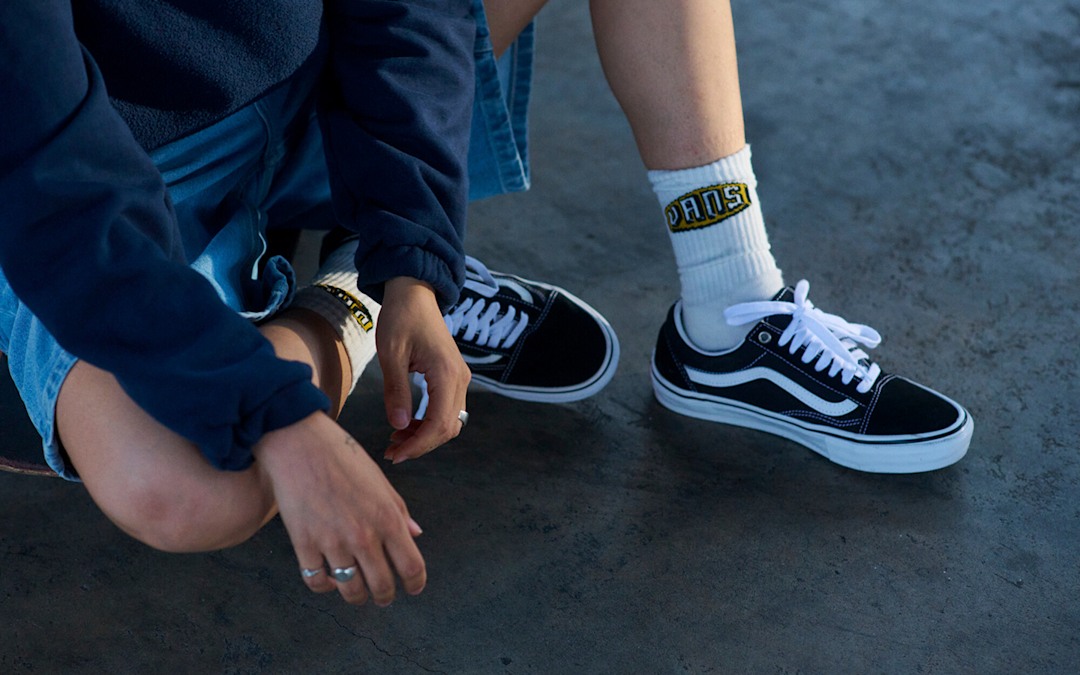 A waist-down shot of a person sitting on a concrete floor wearing denim jeans, white Vans crew socks, and black and white Old Skool.