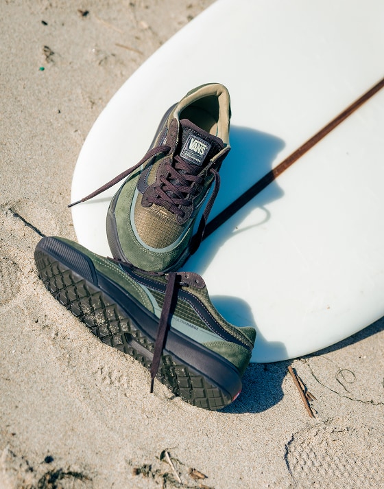 An overhead shot of coal brown UltraRange 2.0 shoes on a surfboard on the beach.