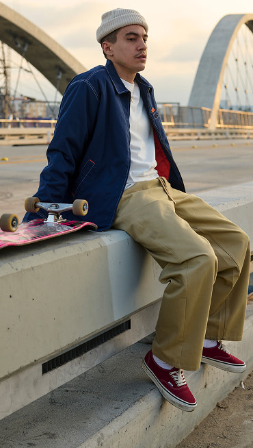 A man sits on a concrete barrier on a bridge wearing brown chinos, a beanie, a white t-shirt, and burgundy Authentic during golden hour.