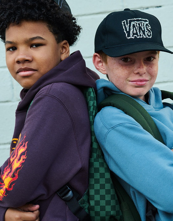 Two boys pose back-to-back with Vans hoodies and backpacks outside of a warehouse.