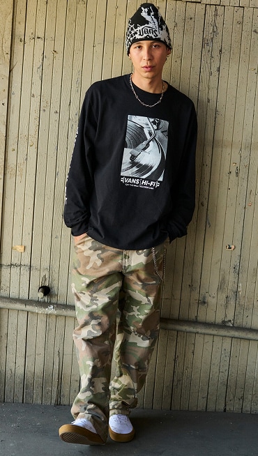 A man poses in white Skate Authentic shoes wearing a black long-sleeve shirt, camo pants, and a beanie outside of a warehouse.