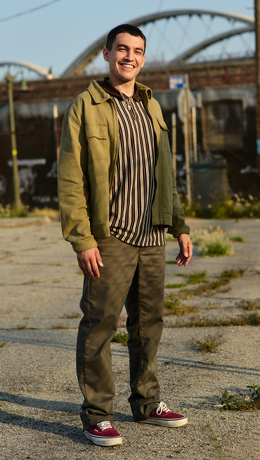 A young man stands in a parking lot wearing a striped shirt, brown chinos, and burgundy Authentic shoes during golden hour.