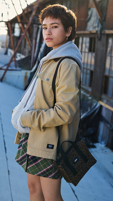 A young woman with short hair and a septum piercing looks at the camera wearing a gray Vans hoodie and a tan Vans jacket.