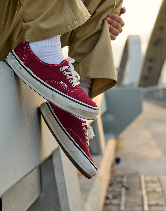 A close-up of a man wearing burgundy Authentic sitting on a concrete barrier with brown chinos during golden hour.
