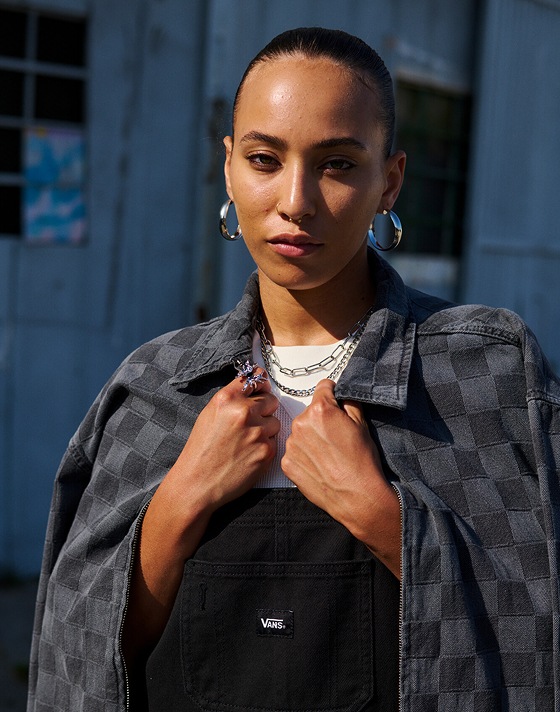 A woman poses on the street wearing black Sirelle Overalls and a gray and black Checkerbord jacket.