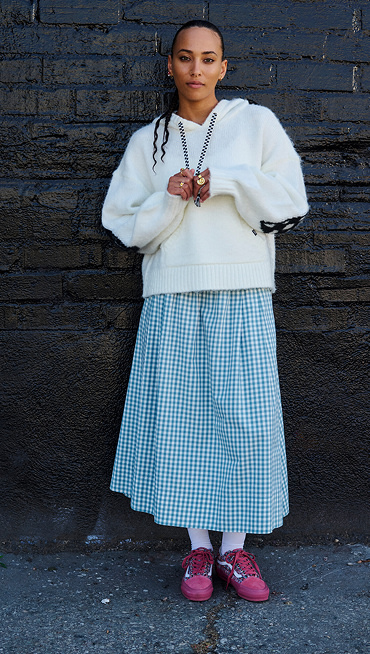 A woman poses against a brick wall for the camera wearing a white hoodie, a light blue gingham dress, and engineered knit Old Skool.