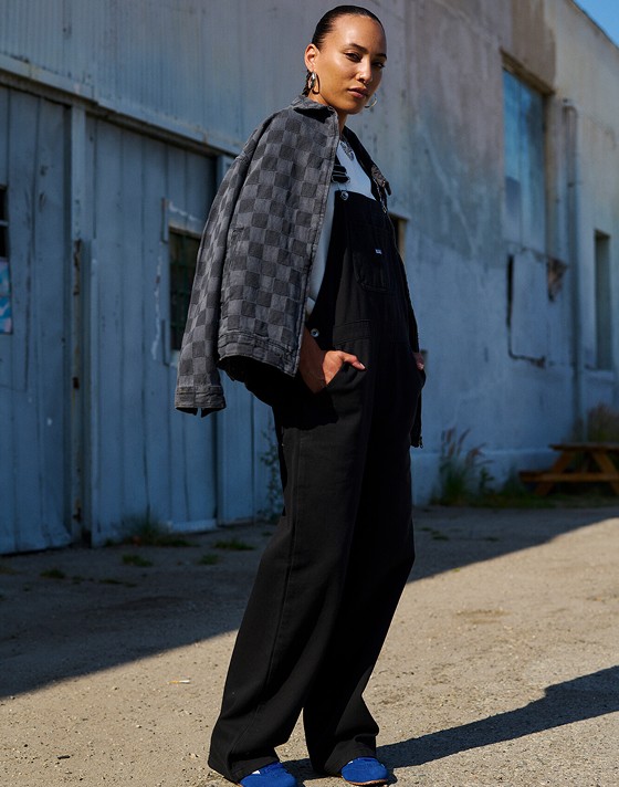 A woman poses on the street wearing black Sirelle Overalls and a gray and black Checkerbord jacket.