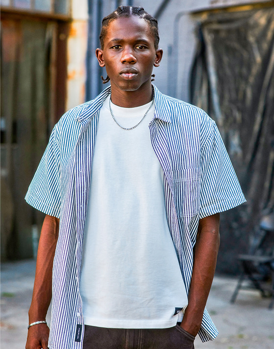 A man looks at the camera wearing a blue and white Lawson Stripe Button Up Shirt, brown jeans, and white t-shirt outside of a warehouse.
