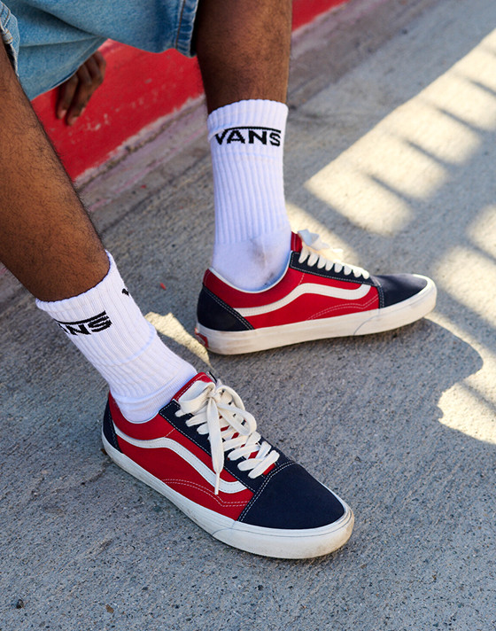 A young man looks down the street and sits on a curb in red and navy Old Skool paired with blue denim jeans.