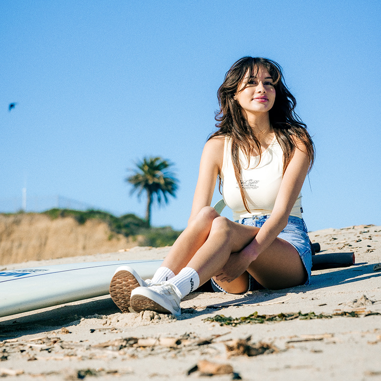 Vans Surf Team Rider Maddie Miller sits on a beach wearing the Everydayness Crop Tank Top and vintage white UltraRange 2.0.