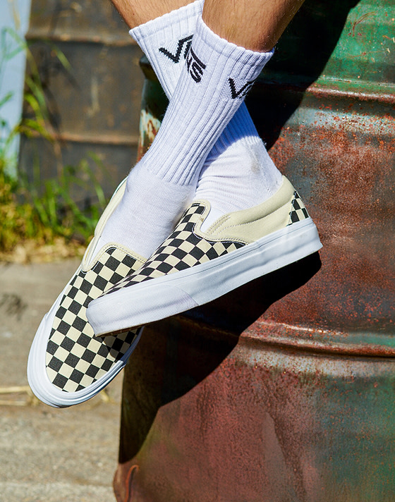 A close-up of a man wearing black and white Checkerboard Classic Slip-On with white Vans crew socks sitting on a rusty oil drum.