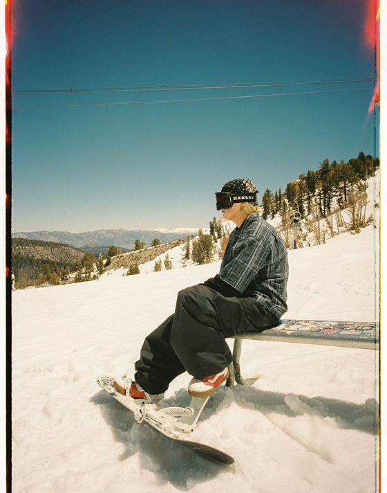 Vans Snowboard Team Rider Mia Brookes wearing red and white Encore Pro snowboard boots and Vans clothes sitting on a metal bench on a ski slope.