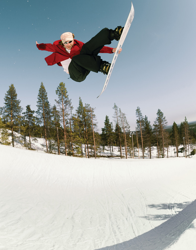 A snowboarder wearing tan snowboard pants and a white sweatshirt grinds a rail on a snow-covered road near a concrete building.