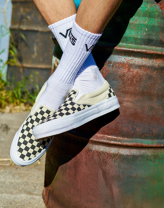A close-up of a man wearing black and white Checkerboard Classic Slip-On with white Vans crew socks sitting on a rusty oil drum.