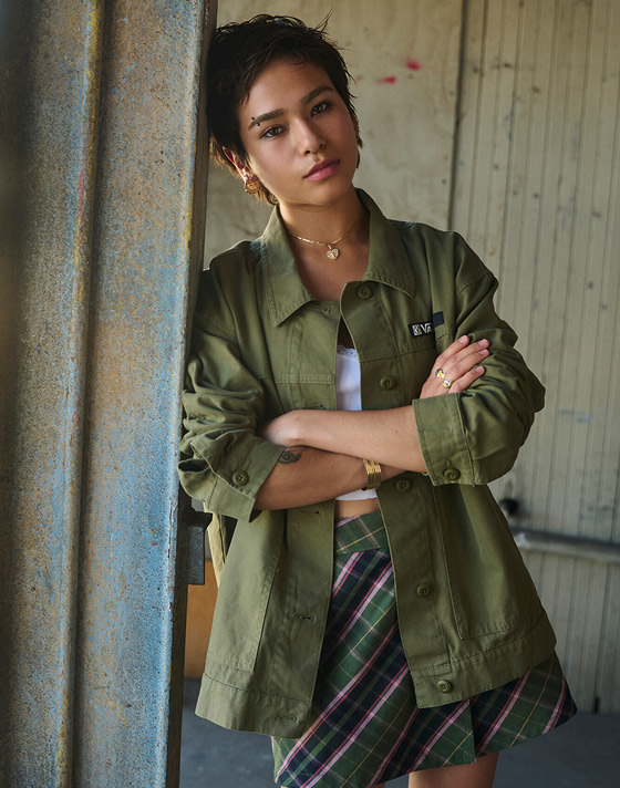 A young woman leans against a wall wearing an olive overshirt and a pine forest green plaid Sirelle Wrap Mini Skort.