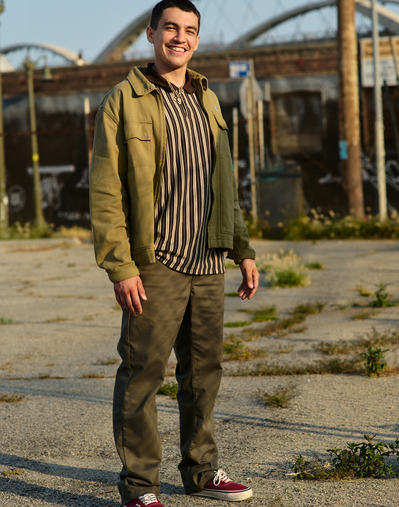 A young man stands in a parking lot wearing a striped shirt, brown chinos, and burgundy Authentic shoes during golden hour.