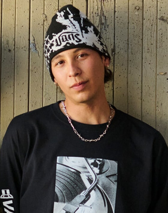 A man in a Vans shirt and beanie looks into the camera with a soft smile.