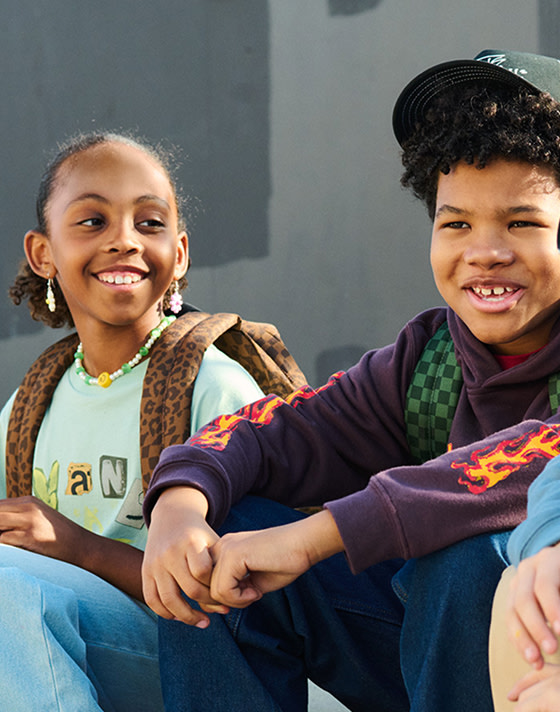 Three young kids sit on a curb wearing Vans clothes, backpacks, Old Skool, Knu Skool, and Checkerboard Classic Slip-On.