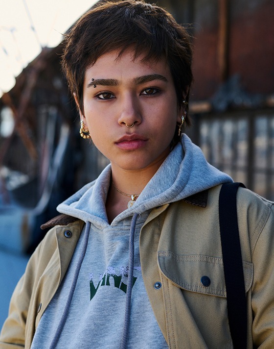 A young woman with short hair and a septum piercing looks at the camera wearing a gray Vans hoodie and a tan Vans jacket.