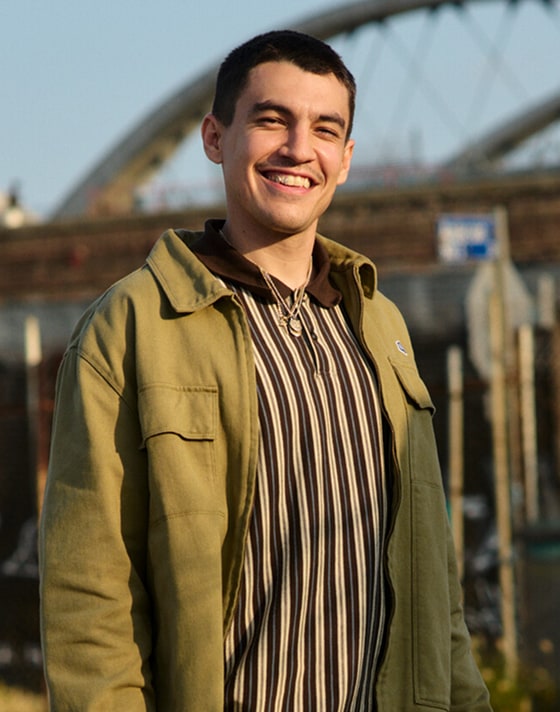 A young man stands in a parking lot wearing a striped shirt, brown chinos, and burgundy Authentic shoes during golden hour.