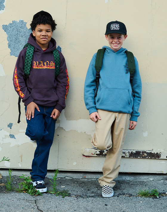 Two kids lean up against a wall wearing Vans clothes, backpacks, Knu Skool, and Checkerboard Classic Slip-On.