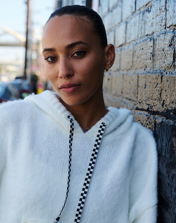 A woman with a septum piercing leans against a brick wall wearing the Kawaii Hoodie looking into the camera.