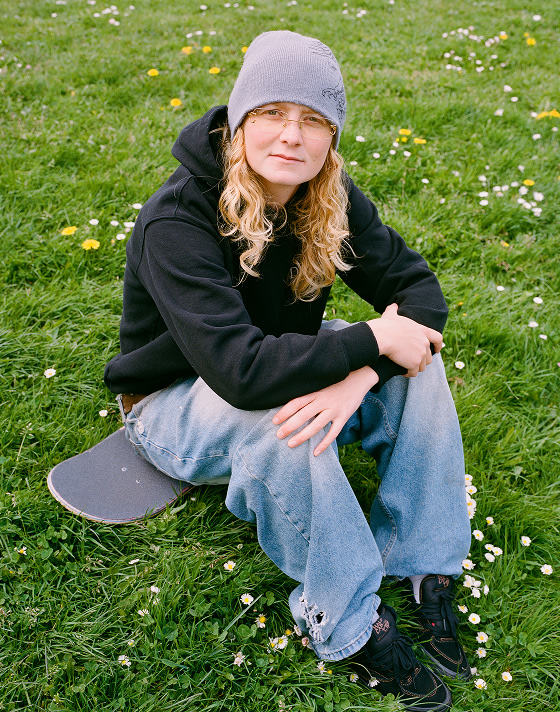 Vans Skate Team Rider Una Farrar looks into the camera and sits on a skateboard in the grass wearing black Skate Half Cab.