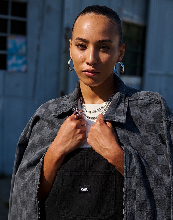 A woman poses on the street wearing black Sirelle Overalls and a gray and black Checkerbord jacket.
