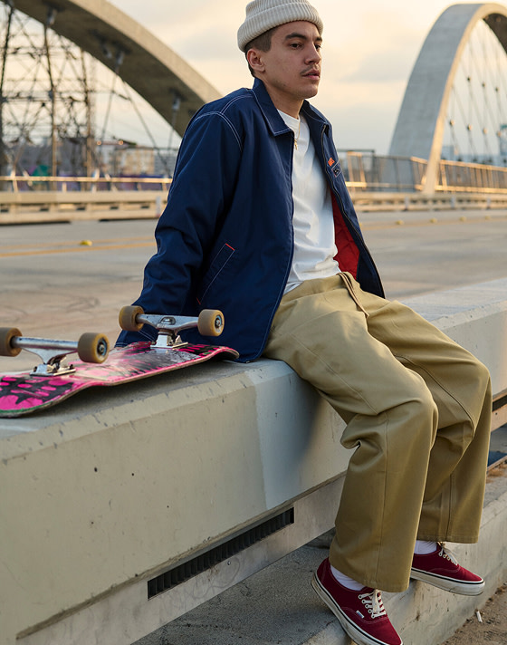 A man sits on a concrete barrier on a bridge wearing brown chinos, a beanie, a white t-shirt, and burgundy Authentic during golden hour.