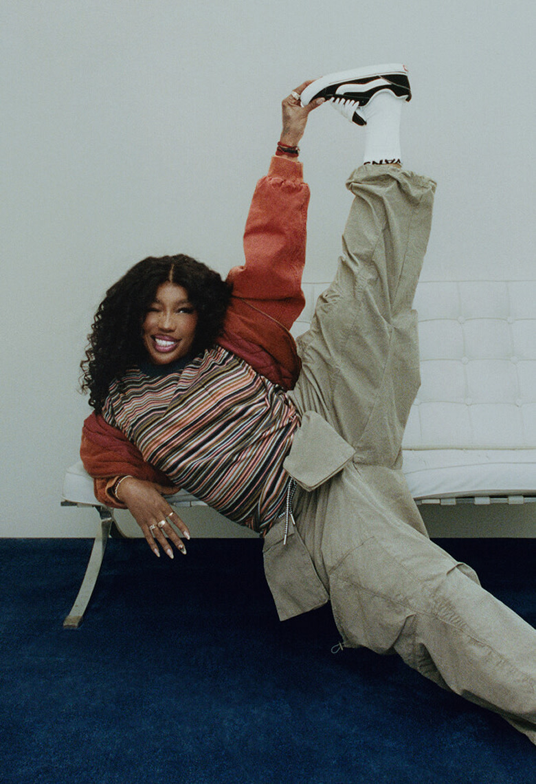 Singer SZA leans on a white sofa with her leg up in the air holding onto her black and white Knu Skool shoe wearing baggy pants and a striped t-shirt.