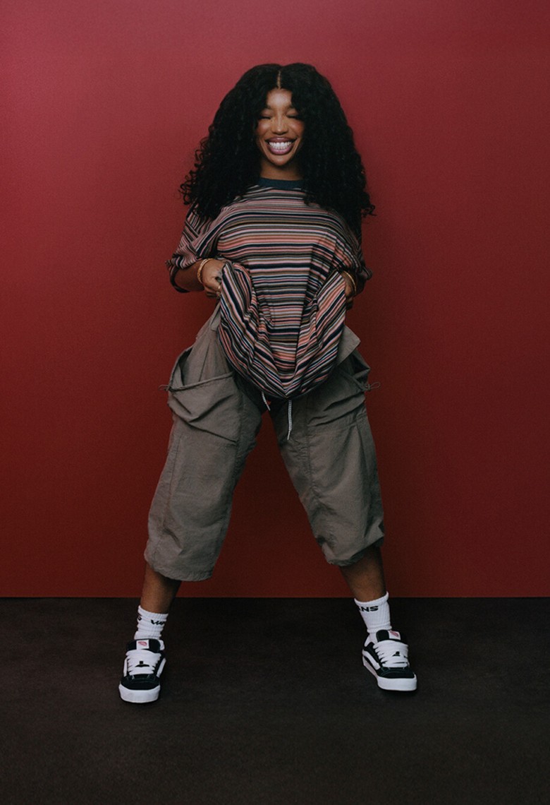 Singer SZA poses in a red room next to a payphone and smiles with her eyes closed wearing a striped t-shirt, and black and white Knu Skool shoes.