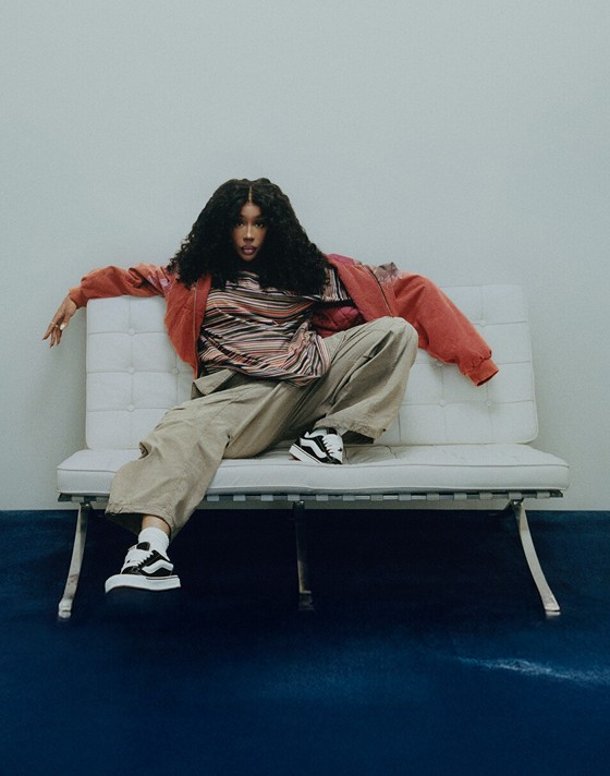 Singer SZA sits on a sofa in baggy pants, a striped t-shirt, and a red zip-up hoodie looking into the camera with black and white Knu Skool shoes.