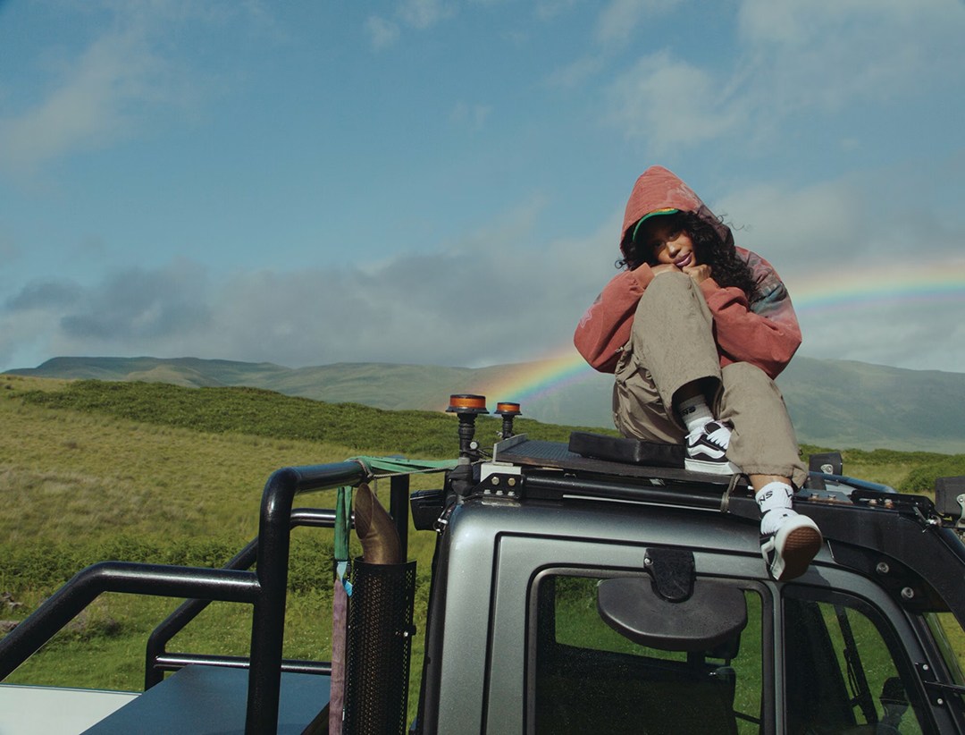 SZA sitting on the roof of a truck, a rainbow can be seen in the background.