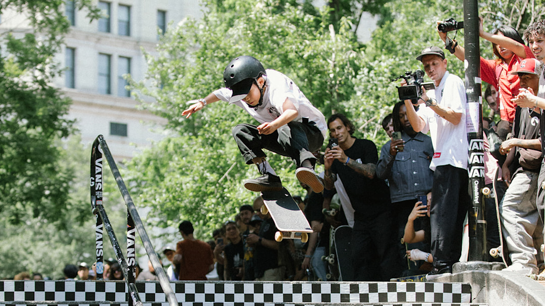 Professional skater Noah Uchida attempting a kickflip on a skateboard down a stair set in front of a crowd.