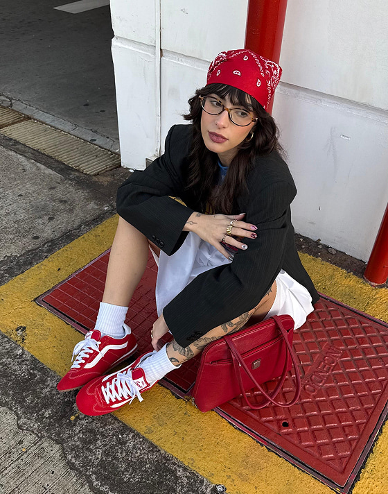 A woman sits on the sidewalk wearing a white skirt, black blazer, and racing red Super Lowpro to match her red bandana and purse.