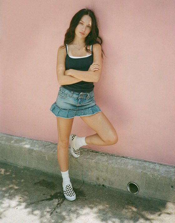 A woman leans on a coral-colored wall wearing a denim skirt, black tanktop, white crew socks, and white and black Checkerboard Classic Slip-On.