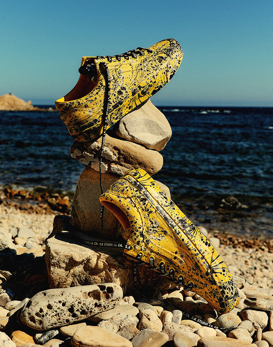A pair of OTW Old Skool 36 Vibram with SRSTUDIOLACA marbled print lie on a beach stacked on rocks with the ocean in the background.