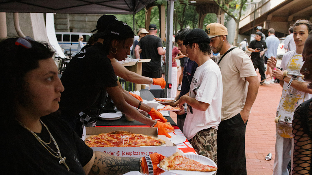 A woman handing out pizza to skateboarders in front of the pizza tent at Go Skate Day 2025 in Brooklyn.