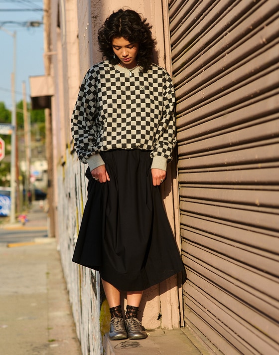 A young woman wearing a black Francesca Full Skirt and the Checkerboard Retro Crew Sweatshirt looks down at her black and silver Super Lowpro.
