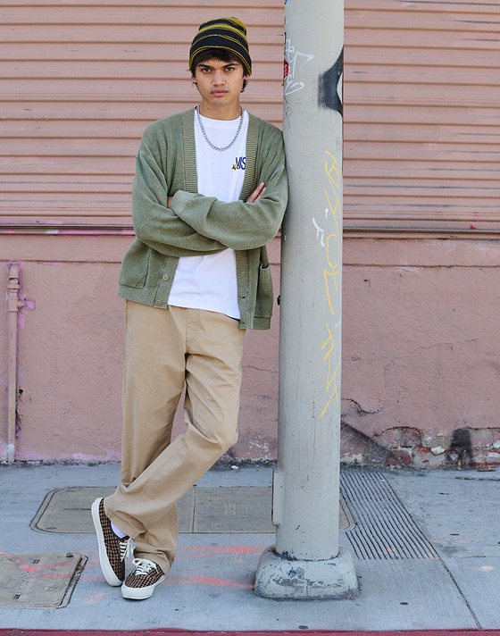 A young man scowls at the camera wearing a white Vans t-shirt and a moss green cardigan as he leans against a metal pole.