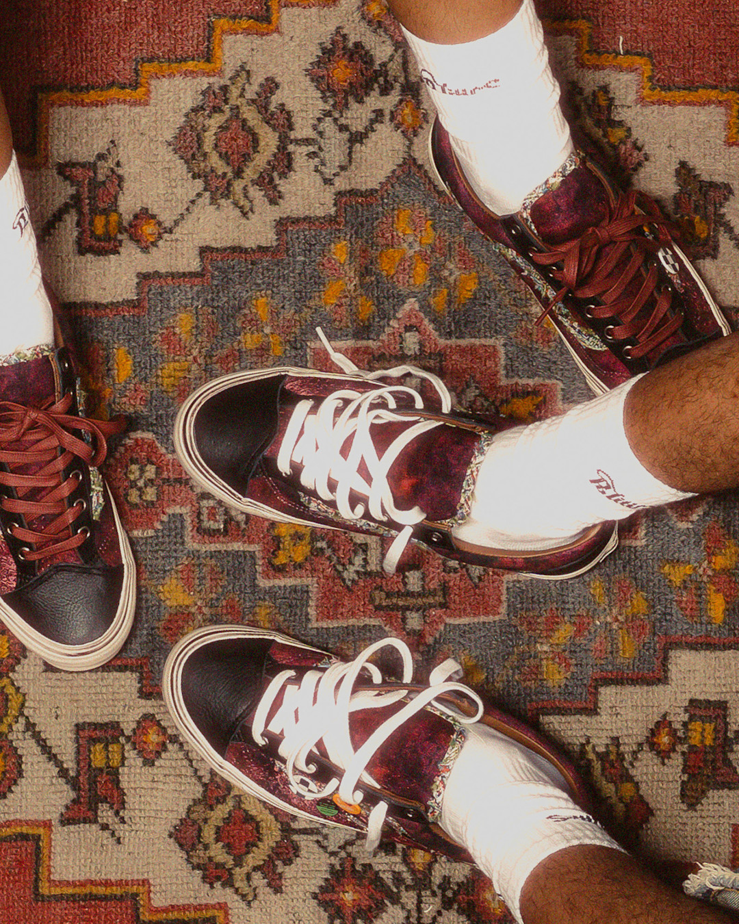 Three pairs of feet wearing matching burgundy Vans sneakers with black toe caps and patterned trim, styled with white crew socks on a patterned rug.