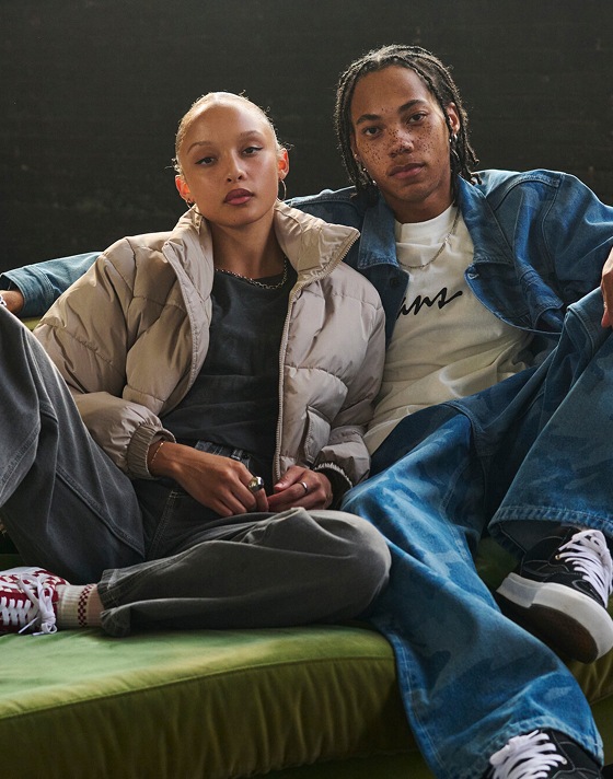 A young man and woman sit on a green felt couch wearing the MTE Hillgate Puffer and a blue camo denim jacket and pants.