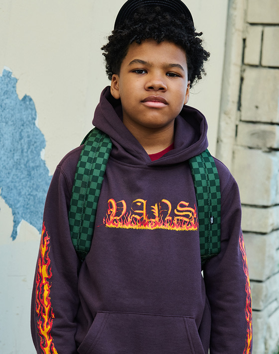 A young boy looks into the camera wearing Kids Vans Inferno Pullover Hoodie and a Kids Old Skool Grom Backpack.