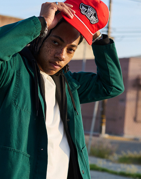 A man looks into the camera fixing his red Vans “Off the Wall” hat paired with a green overshirt.