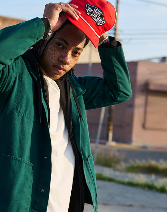 A man looks into the camera fixing his red Vans “Off the Wall” hat paired with a green overshirt.