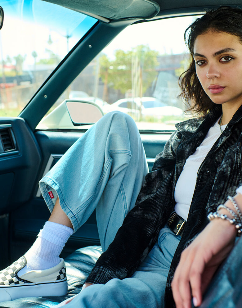 A woman sits shotgun in an old Chevy wearing Check-5 Baggy Denim Pants, a corduroy black overshirt, and Checkerboard Classic Slip-On.