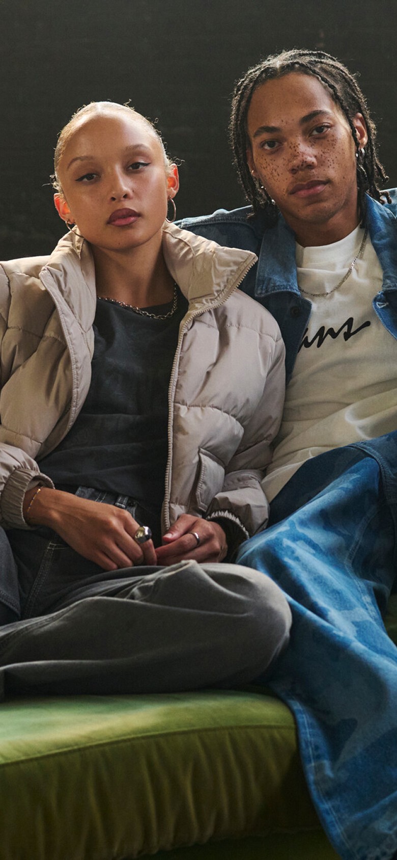 A young man and woman sit on a green felt couch wearing the MTE Hillgate Puffer and a blue camo denim jacket and pants.